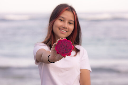 Portrait of girl holding half a dragon fruit in front of her. Selected focus on red dragon fruit. Blurred smiling face.