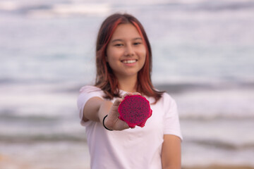 Portrait of girl holding half a dragon fruit in front of her. Selected focus on red dragon fruit. Blurred smiling face.