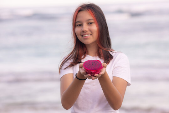 Portrait of girl holding half a dragon fruit in front of her. Selected focus on red dragon fruit. Blurred smiling face.