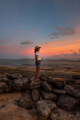 Slim teenager girl standing on a stone on the beach. Amazing sunset background. Colorful sky. Profile face. Ocean view