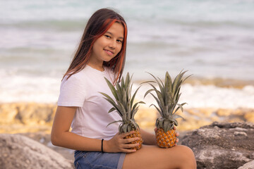 Beautiful teenager girl sitting on the stone, holding two whole pineapples. Smiling face. Organic fruit concept. Bali.