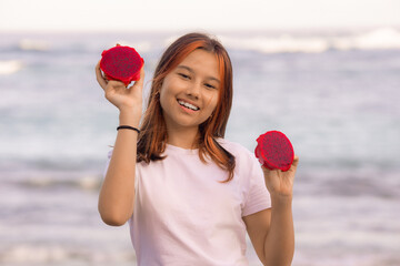 Portrait of happy Asian teenager girl holding red dragon fruit halves. Outdoor photoshoot with dragon fruits. Bali.