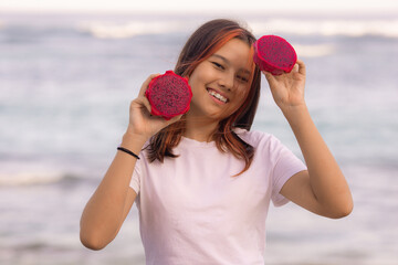 Portrait of beautiful Asian teenager girl holding red dragon fruit halves. Outdoor photoshoot with dragon fruits. Bali.