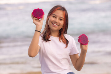 Portrait of charming Asian teenager girl holding red dragon fruit halves. Outdoor photoshoot with dragon fruits. Bali.