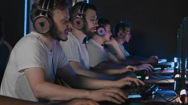Male players are constantly talking through professional gaming headset. Five guys are using gaming mouse keyboard while playing first-person shooter game. LAN games cyber sport tournament