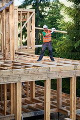 Carpenter building wooden frame house near the forest. Bearded man with spectacles holding large board on his shoulder, dressed in work clothes and helmet. Concept of modern, sustainable construction.