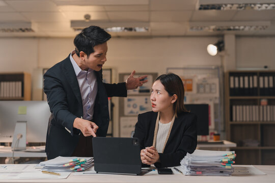 Asian businessman pointing at laptop and scolding female intern for bad work results while working late in office at night, concept of business conflict