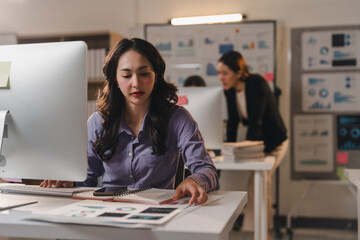 Young asian businesswoman reviewing financial reports and analyzing marketing data while working late at night in a modern office, demonstrating dedication and professionalism