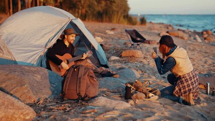 Male tourist playing guitar in tent and woman filming his by smartphone. Travelers spending weekend trip in nature, camping on seashore in summer day, travel bloggers enjoying music and relaxation