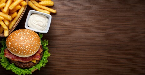 Fototapeta premium Overhead shot of a cheeseburger with bacon and lettuce, served with a basket of golden french fries and a small bowl of mayonnaise, all arranged on a dark brown wooden background. 
