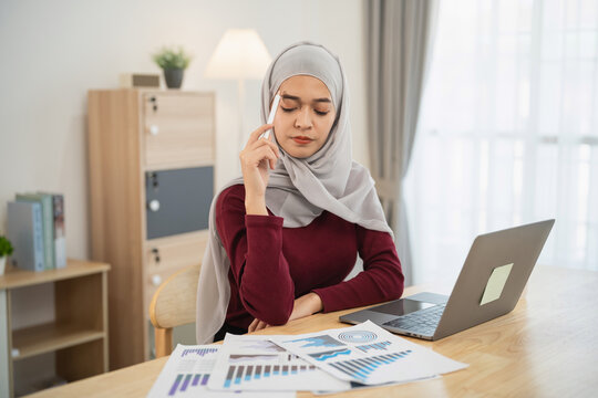 Thoughtful young woman in hijab analyzing financial data at her desk with laptop and charts, representing business strategy and decision-making - Powered by Adobe