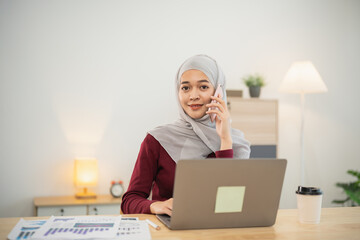 Professional young woman in hijab working on laptop while talking on phone in modern office environment