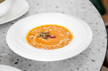 A bowl of creamy orange soup garnished with seeds and nuts sits on a textured grey table. Perfect for showcasing healthy eating and culinary arts.
