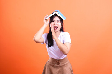 A woman with a book on her head and wearing a white shirt poses in front of orange background