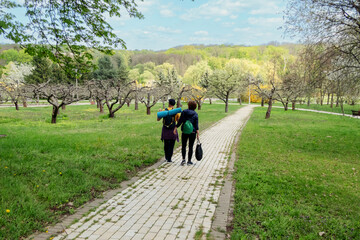 Fototapeta premium Two people walking on a paved path in a lush park with blooming trees, ideal for nature, outdoor activities, and springtime themes.