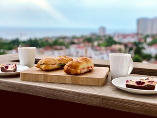 Outdoor breakfast scene. Close up of a wooden table with sandwiches, cheesecake, and coffee with a beautiful deffocused urban background, creating a relaxing setting for enjoying a meal outdoors.