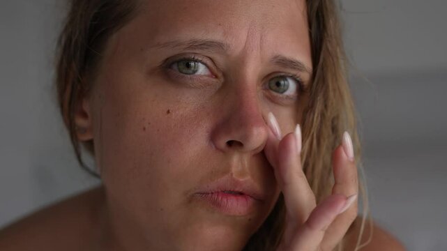 Close-up of a young blonde woman gently touching her sunburned nose, which is covered with moisturizing cream. Her expression is tender and careful, showing skincare and sun damage relief
