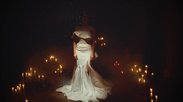Fantasy woman queen in luxurious long white dress walking under falling snow, dark room full many candles burning. sexy fashion model girl princess enjoy winter magic New Year holiday. fairy tale elf