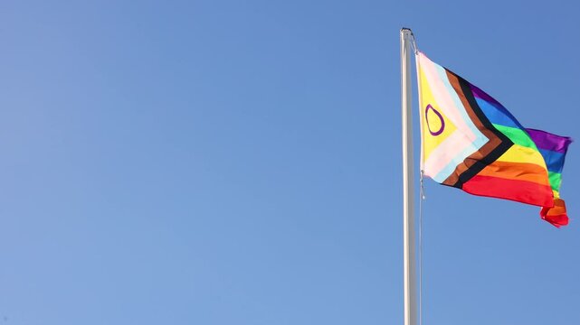 Intersex-inclusive redesign of the Progress Pride Flag. Rainbow LGBT flag against a blue sky. Flag fluttering in the wind. An abbreviation that stands for lesbian, gay, bisexual, transgender