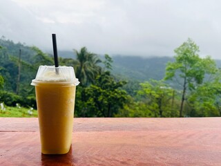A disposable cup with a straw sits on a rustic wooden podium, surrounded by lush greenery and rolling tropical hills in the background. 