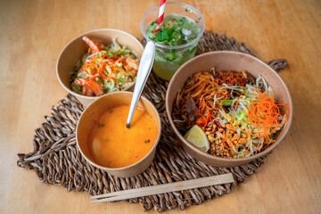 Coconut soup, pad thai, vegetable salade with shrimp in bowl and cup of cucumber lemonade with straw on bamboo pad.