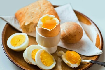 Boiled egg in egg cup, in shell and sliced on plate, spoon and bread (bun), towel.