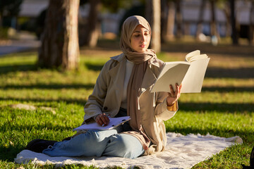 Young muslim woman studying in a park, taking notes from a book