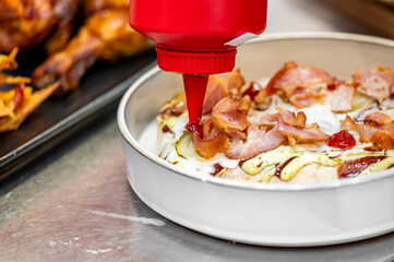 A close-up of a delicious pizza being topped with ketchup and bacon. The scene captures the savory ingredients being drizzled onto the pizza, emphasizing a casual dining experience.
