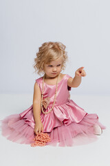 Cute girl in pink dress playing with beads in a bright studio setting, expressing curiosity with a pointed finger