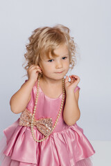 Little girl in pink dress with curly hair posing with pearl necklace against a plain background