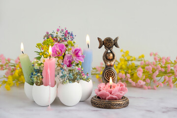 amulet of wiccan Goddess figurine, floral candle and candles with flowers in eggs shells on light background. Symbol of the Triune Moon. esoteric ritual for Ostara, spring season. witchcraft