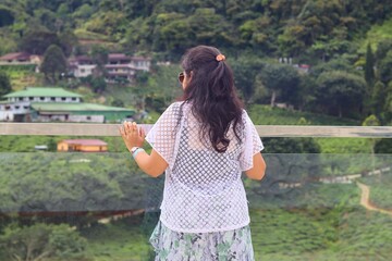 S cenic viewpoint of Cameron Highlands A woman with long, dark hair, wearing a white lace top and a floral skirt, standing with her back to the camera while overlooking a lush, green landscape.