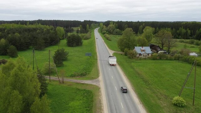 Aerial view of a truck transporting a modular home on a countryside highway, surrounded by green fields, trees, and passing cars.