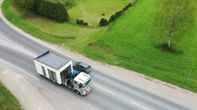  Aerial view of a truck transporting a modular home on a curved rural road, passing a green landscape with a farmhouse and a small car.