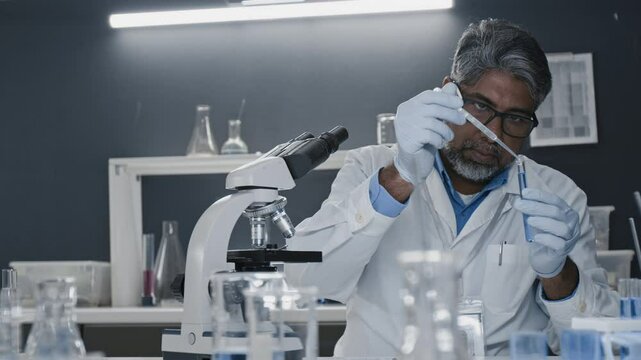 Medium shot of senior Indian biologist in lab coat adding drops of reagent into solution in test tube and working with microscope, workday in breakthrough drug development laboratory