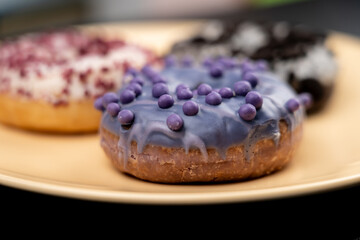 Donuts, macro shot.
Three donuts are lying on a yellow plate.
The focus is on a purple donut with glossy glaze and small round sprinkles.