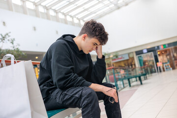 Tired teenage boy with shopping bag sitting alone in mall, exhausted adolescent taking break from...