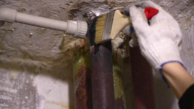 Construction worker wearing protective gloves carefully applying black bituminous paint on metal pipes to prevent corrosion and extend their lifespan