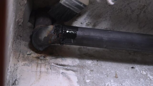 Construction worker wearing protective gloves carefully applying black bituminous paint on metal pipes to prevent corrosion and extend their lifespan