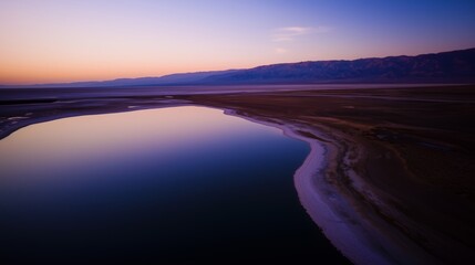 Calm lake with reflective water in arid desert landscape at sunset. Distant mountains under purple sky with horizon glow. Serene nature scene with vast open terrain.