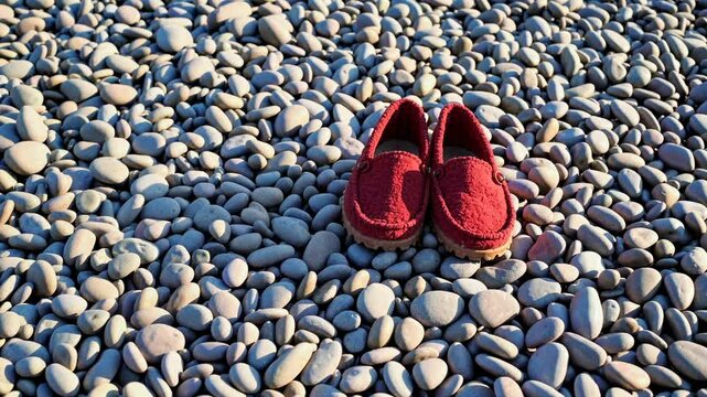 Bright red slippers sit alone on a smooth pebble beach. The sun illuminates the scene, highlighting the variety of rounded stones that create a calming coastal atmosphere
