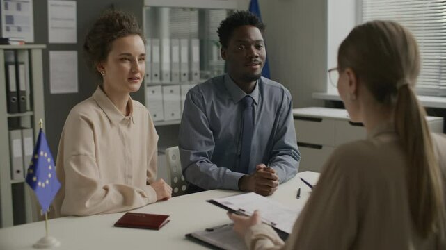 Young diverse couple smiling and answering questions from female consular officer while attending immigration interview at European Union embassy