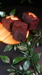 Tempting premium brownies, dark chocolate in color with crunchy almond topping slices, showing the details and texture of the soft cake, looks elegant with artificial leaves and a dark background.
