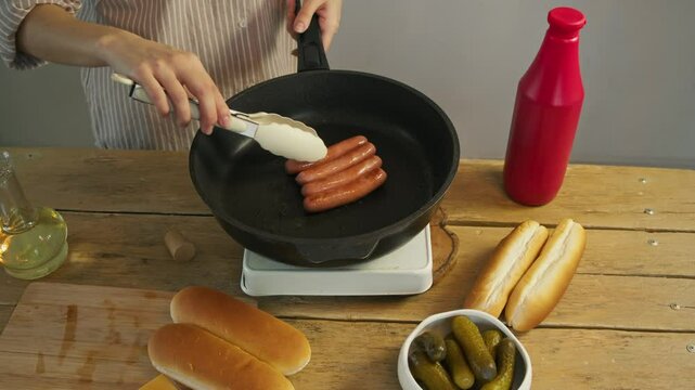The chef makes sure that the sausages, prepared from meat, do not become overcooked by turning them over while they are being fried in a pan, using tongs.
