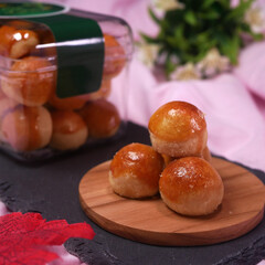 Pineapple-filled nastar cookies in a clear plastic jar with green decoration. Some cookies are served on a wooden board, surrounded by red leaves and pink fabric.