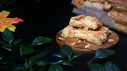 Crispy and delicious cheese Kastengel cookies typical of Eid on a wooden stand and in a plastic jar, decorated with green leaves on a black table, look elegant and premium.