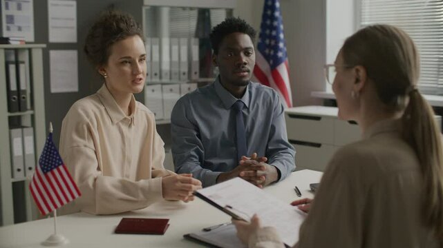 Young diverse couple handing application form to female consular officer, smiling and speaking with her during visa interview at US embassy with American flags