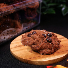 Crispy chocolate cookies with choco chips on a wooden plate, a cookie-filled jar in the background, greenery, and a warm atmosphere, perfect for Eid treats.