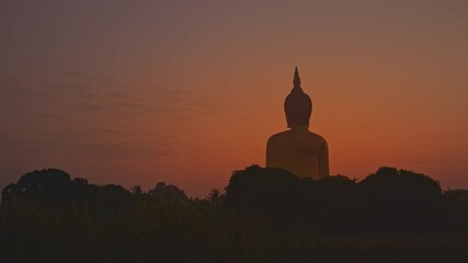 A serene scene featuring a large Buddha statue silhouetted against a golden sunrise.The peaceful rural landscape and soft morning light create a tranquil and spiritual atmosphere. Wat Muang Ang Thong