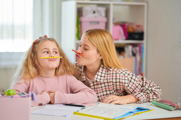 Fototapeta premium Mother and daughter joke around while studying, placing pens under noses, enjoying the moment. Happy home learning, playful approach to schoolwork